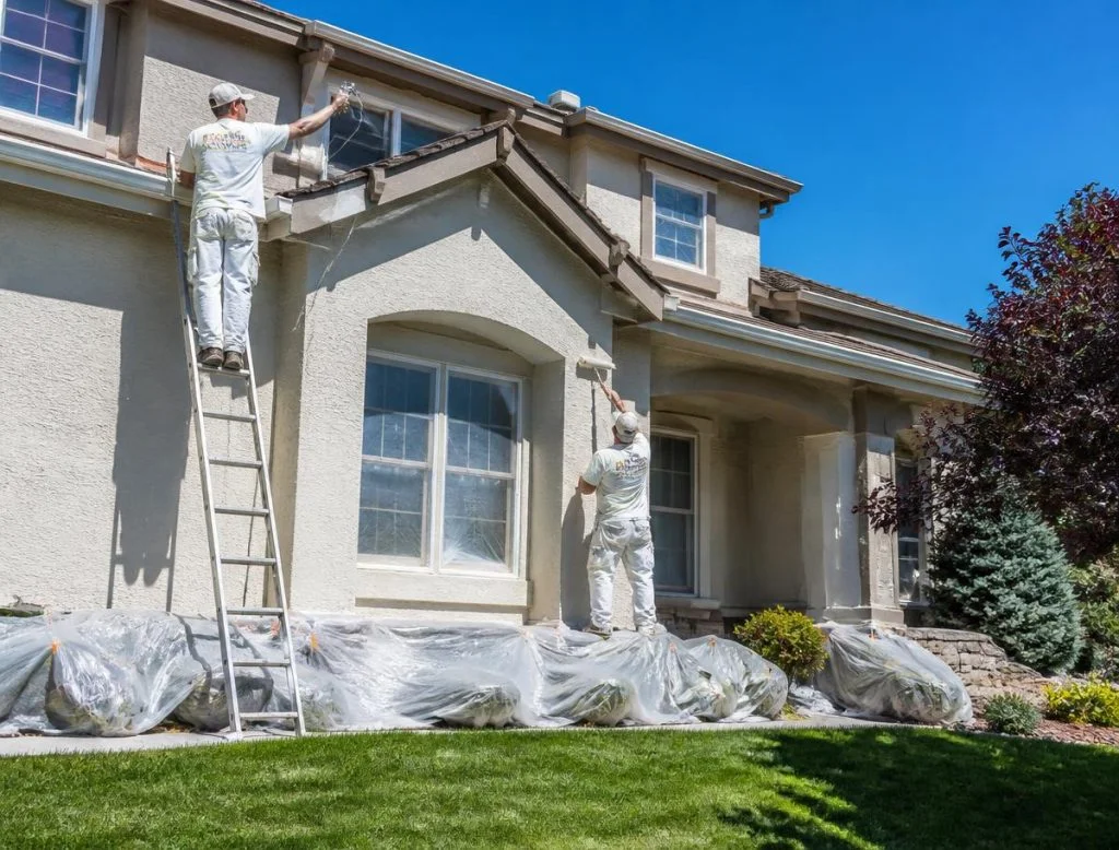 Painters on ladders applying exterior paint to stucco house with protective coverings on landscaping