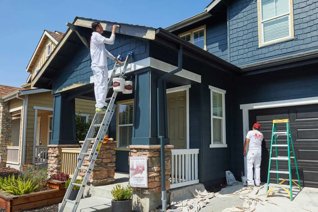 Painters applying dark blue exterior paint to house siding with white trim and stone base